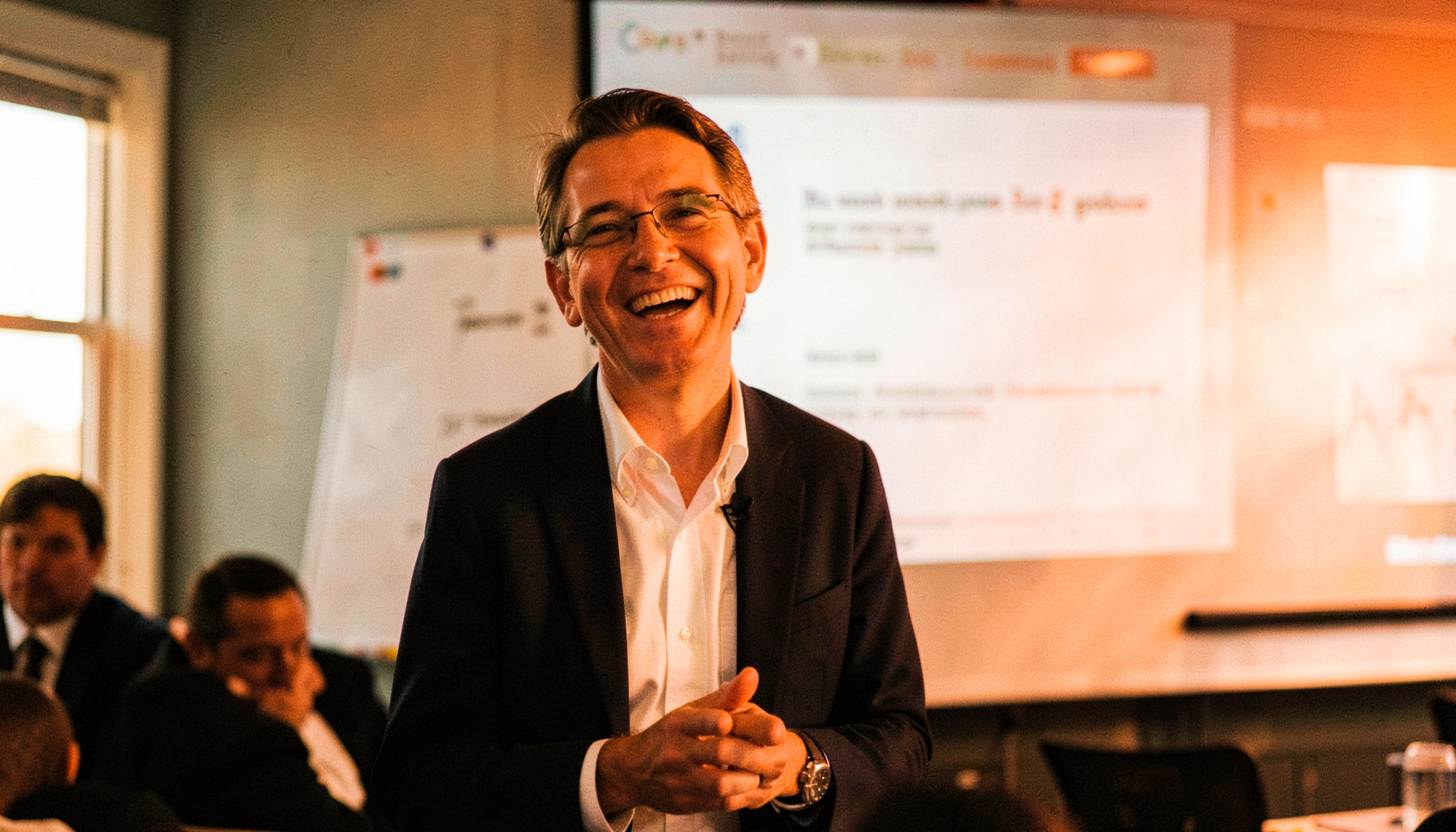 A smiling professional speaking at a staff training session, presentation slides visible on a screen behind them